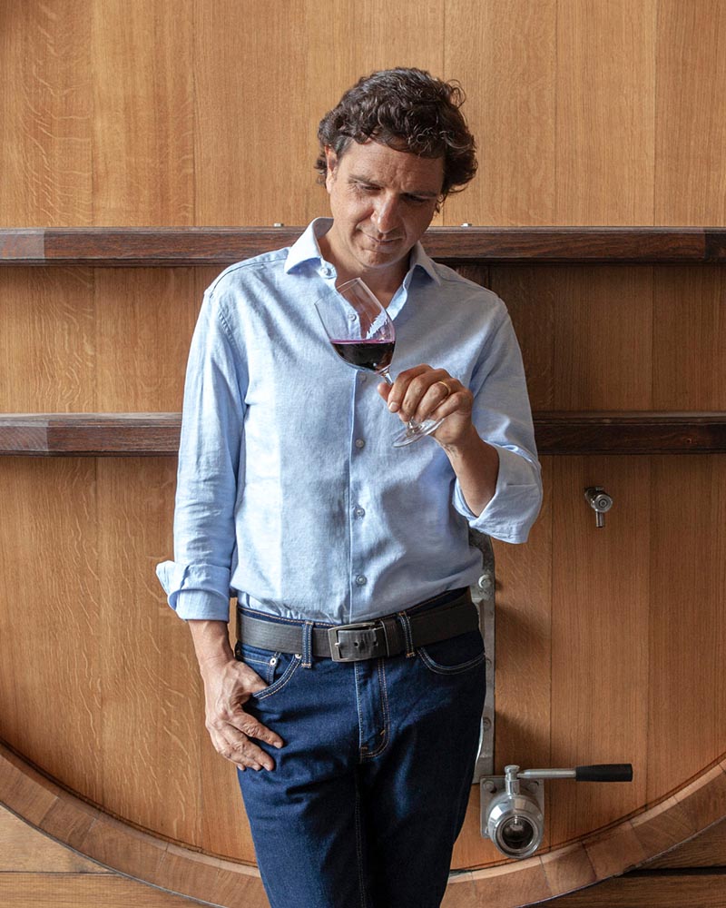 A bio photo of Matías Ríos, standing in front of a wine barrel, examining the wine in a glass. 