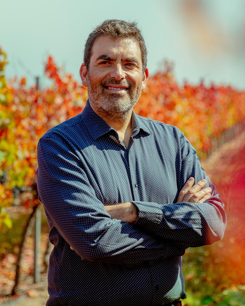 A smiling bio photo of Marcio Ramírez with a blurred, autumnal coloured vineyard in the background. 