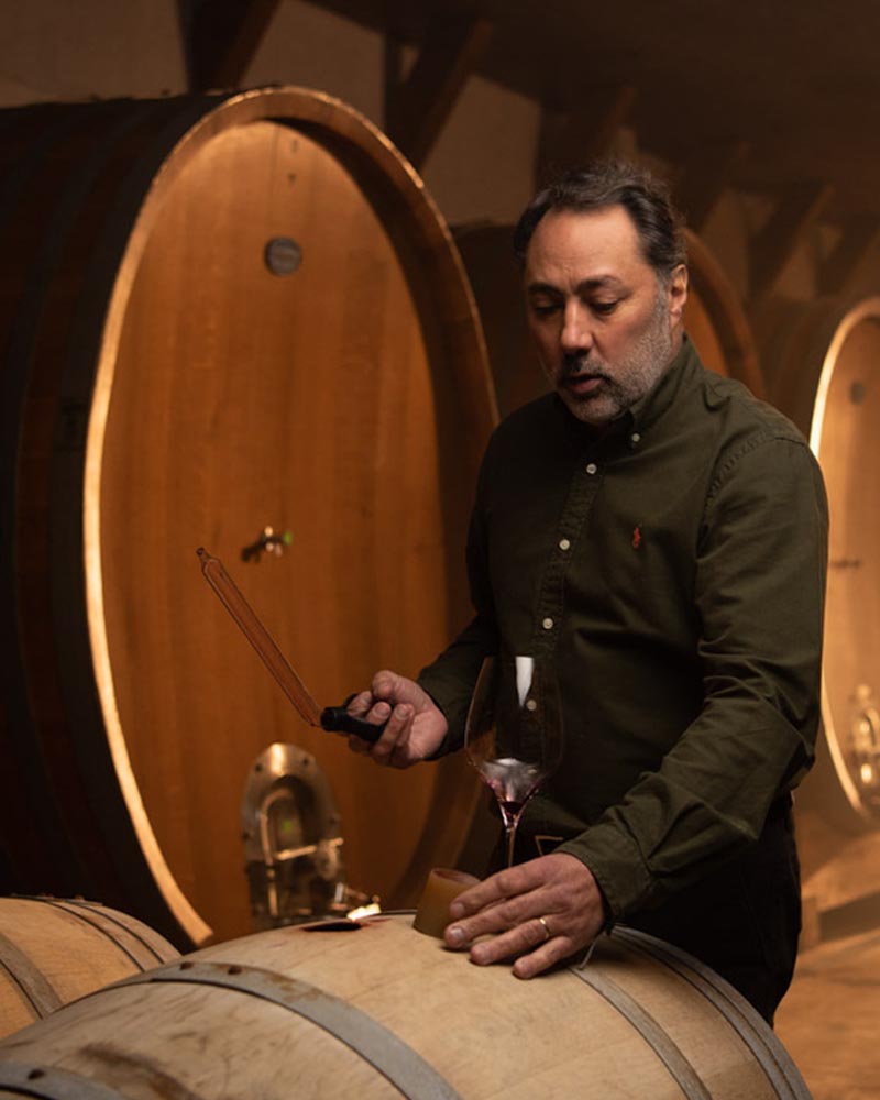 A bio photo of Marcelo Papa tapping a wine barrel in the winery with barrels lined behind him. 