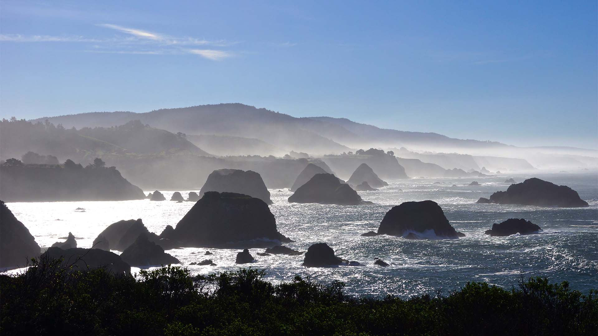 Scenic view of a Californian coast with dark bushes in the foreground, jagged rocks jutting out of the sea in the mid-ground, and a mountain range with a clear blue sky in the background. 