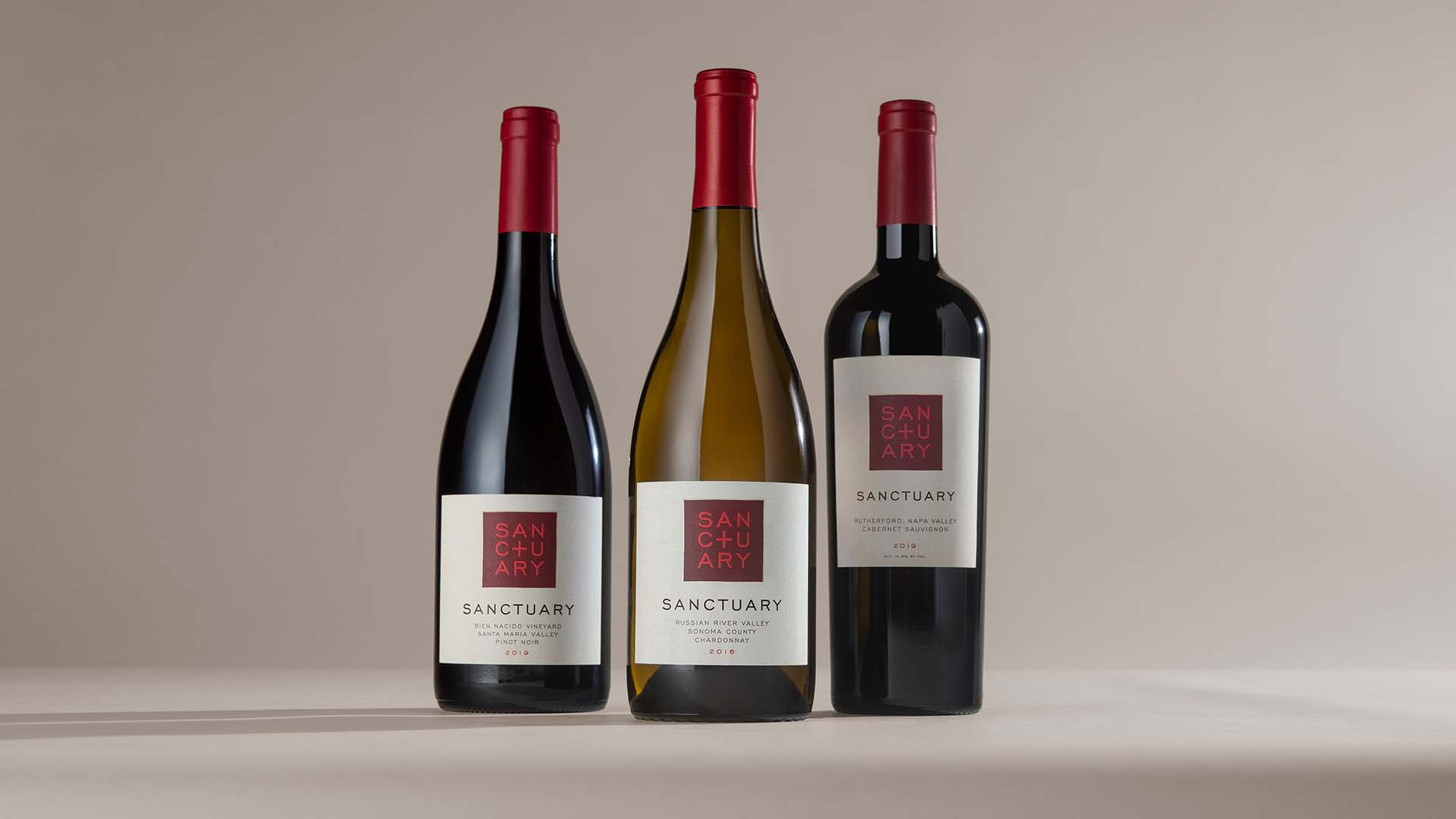 A bottle of Sanctuary's pinot noir, chardonnay, and cabernet sauvignon lined up and centred on a beige backdrop. 