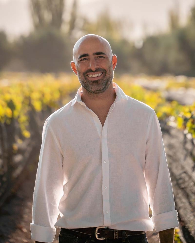 A smiling bio photo of Germán Di Césare standing between a row of green vines, blurred in the background. 