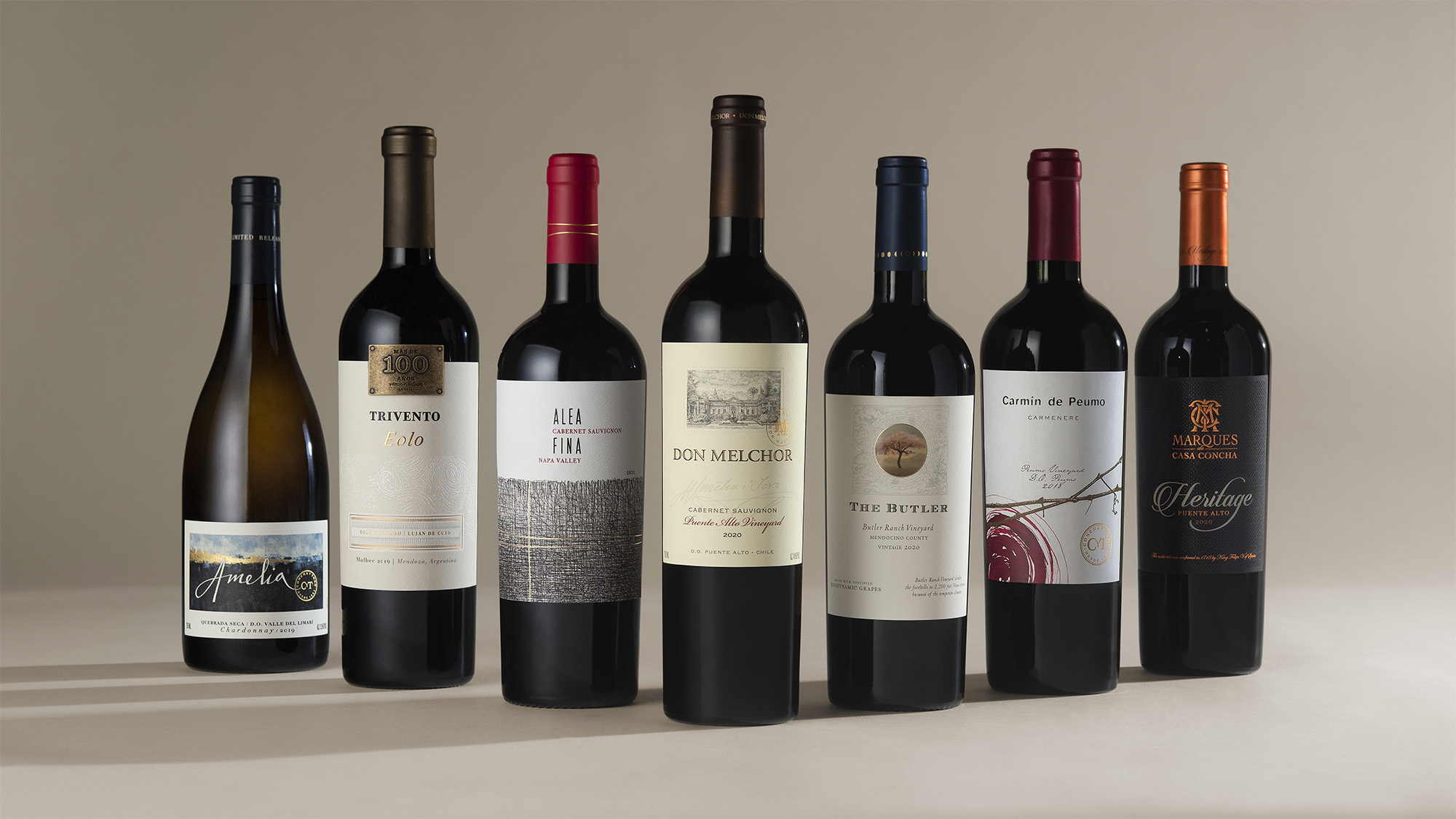A studio photograph of seven bottles of wine from various vineyards positioned in a line. The studio background is a cool beige. 