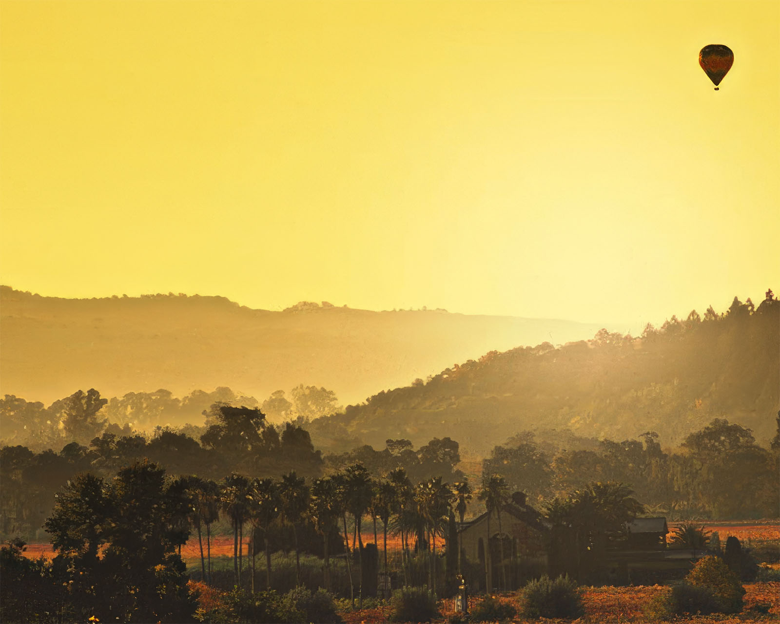 A photograph of a Napa Valley landscape covered in a sunset yellow haze with palm trees and a silhouette of a house in the foreground and two tree-covered mountains in the background.  